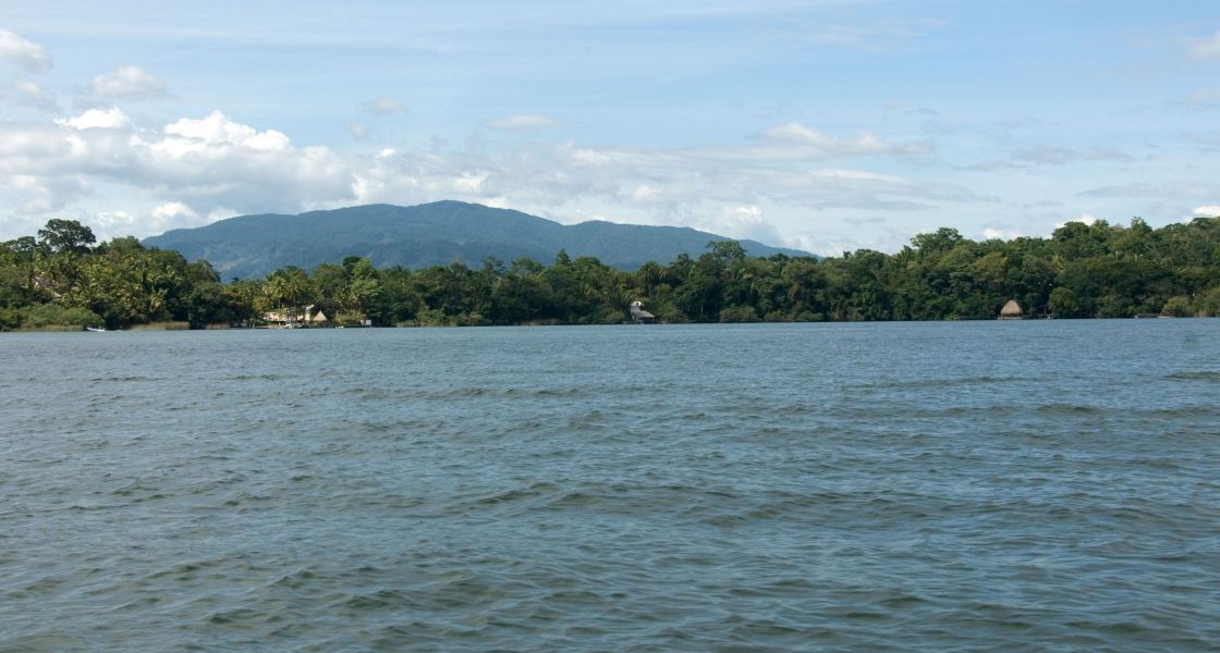 Ein See mit Bäumen am Ufer und im Hintergrund ein hoher Berg.