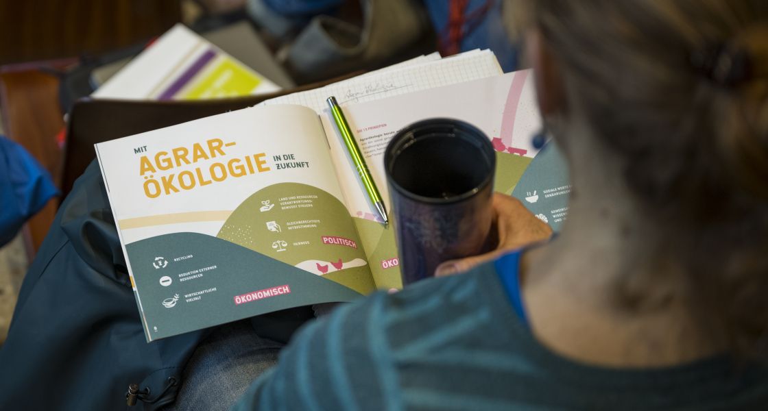 Die Broschüre Agrarökologie liegt offen auf dem Schoß einer Person in einem Hörsaal