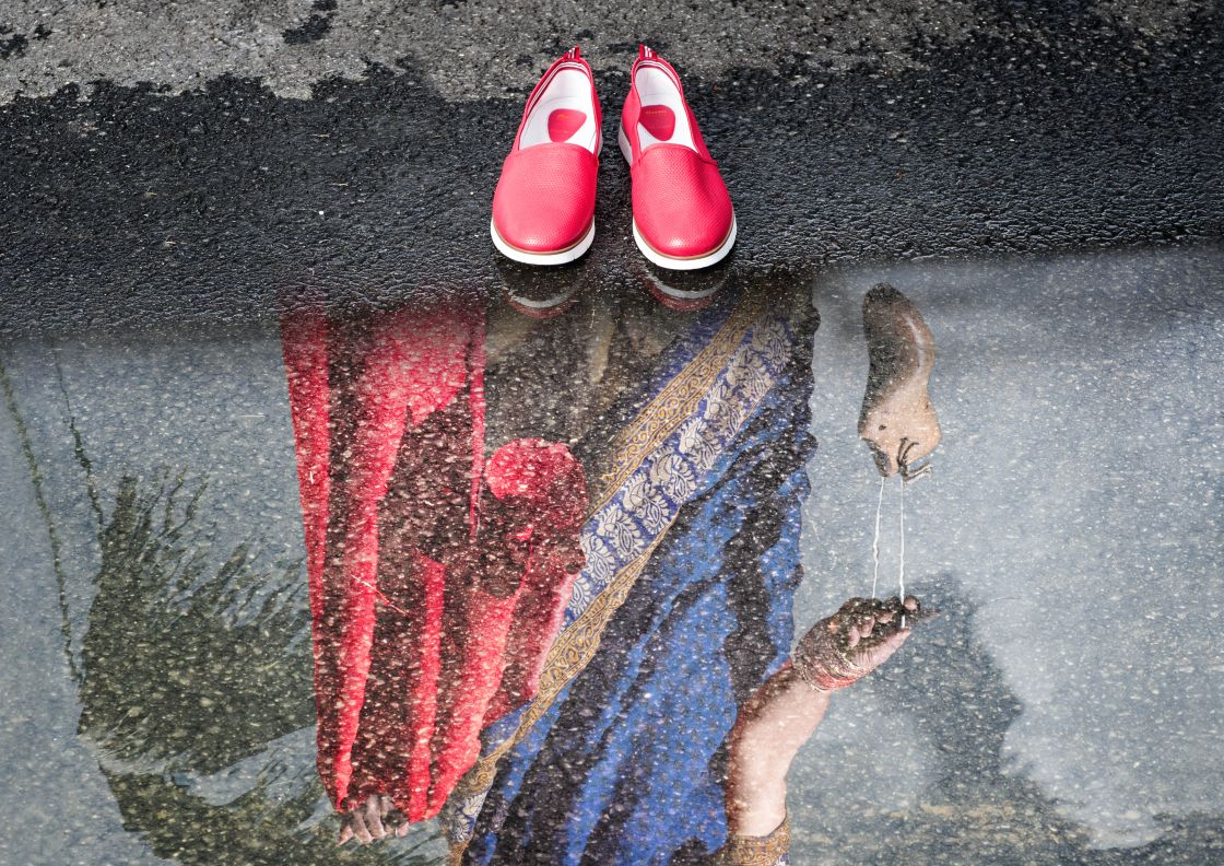 ein paar rote flache Schuhe stehen vor einer Pfütze und man sieht das Spiegelbild einer Frau
