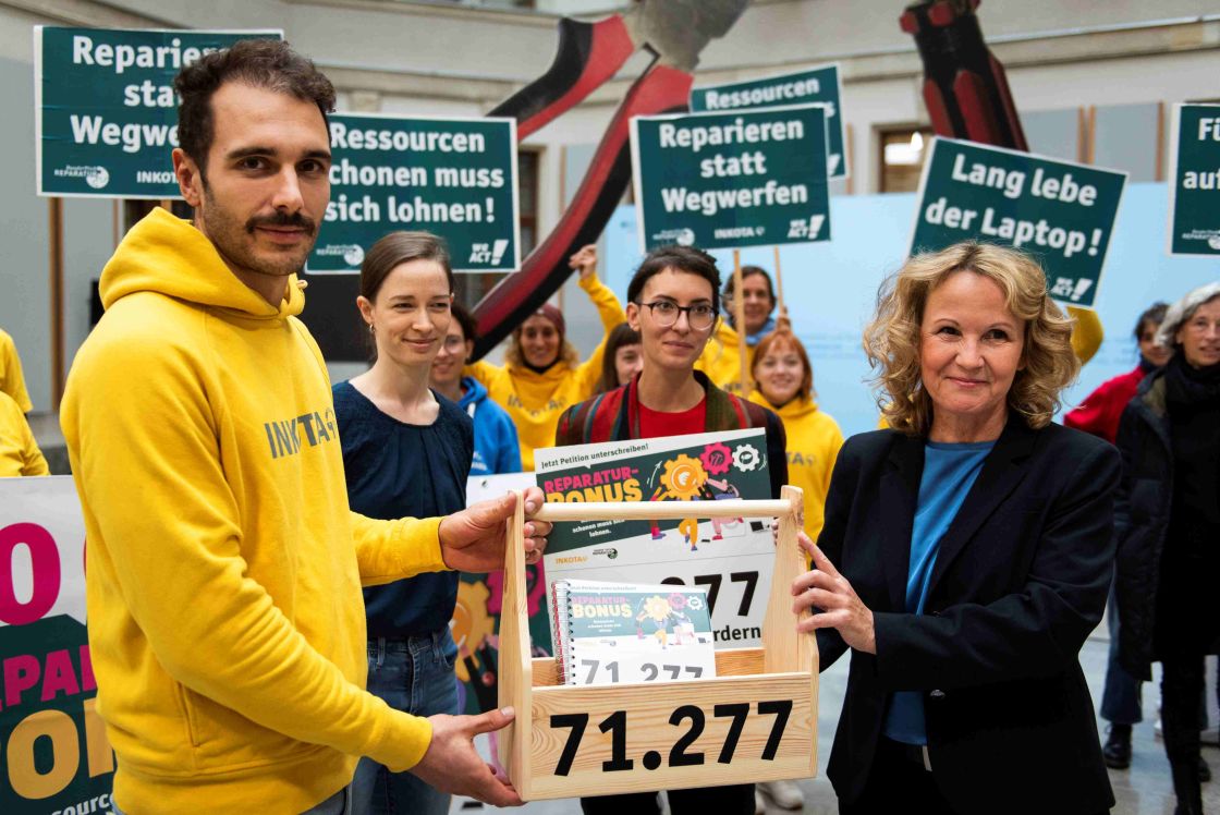 Ein Mann und eine Frau halten gemeinsam einen Holzkasten fest, auf dem die Anzahl der Unterschriften für eine Petition geschrieben steht