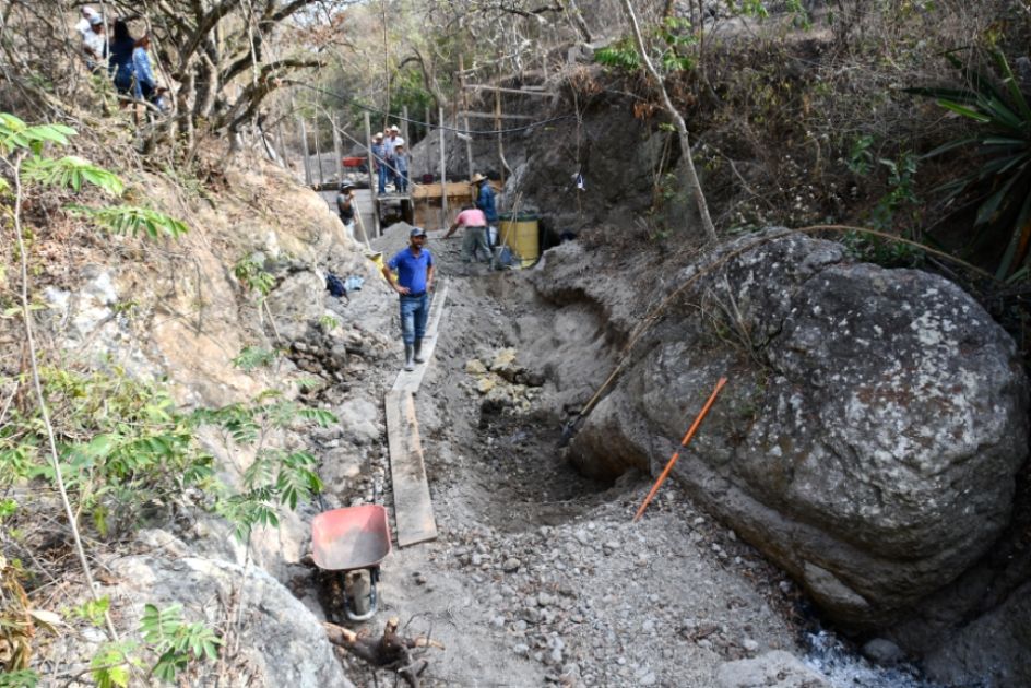 Ein ausgetrocknetes Flussbett, im Vordergrund ein großer Steinblock, Bretter, einige Arbeiter, Schubkar