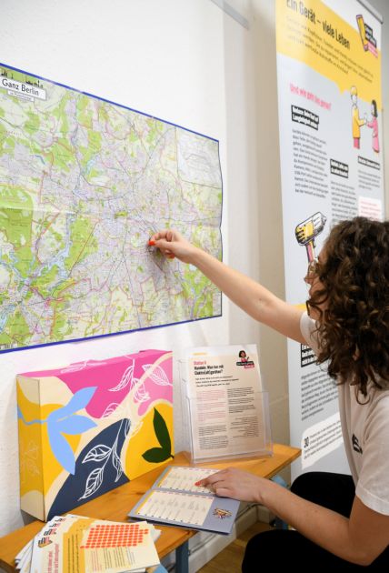 Weiblich gelesene Person klebt an einer Ausstellungsstation rote Punkte au einen Stadtplan von Berlin