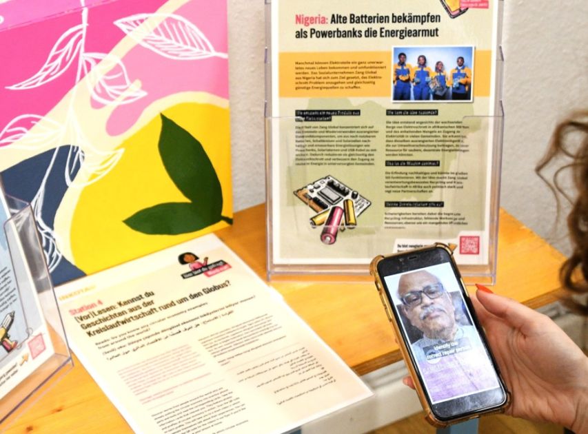 Eine Person schaut ein Video über Kreislaufwirtschaft auf dem Handy, von der Person ist nur die Hand zu sehen, die das Handy hält und ein Teil vom Hinterkopf. Auf einem Tisch liegen Infoblätter mit QR-Codes zum einscannen. 