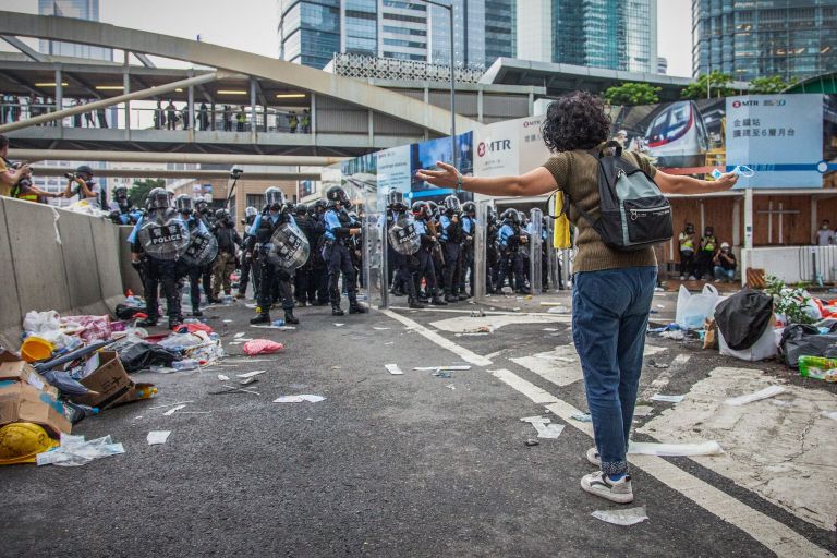 Eine Frau steht auf einer urbanen Straße, ihr gegenüber stehen Polizist*innen. | Studio Incendo (CC BY 2.0)