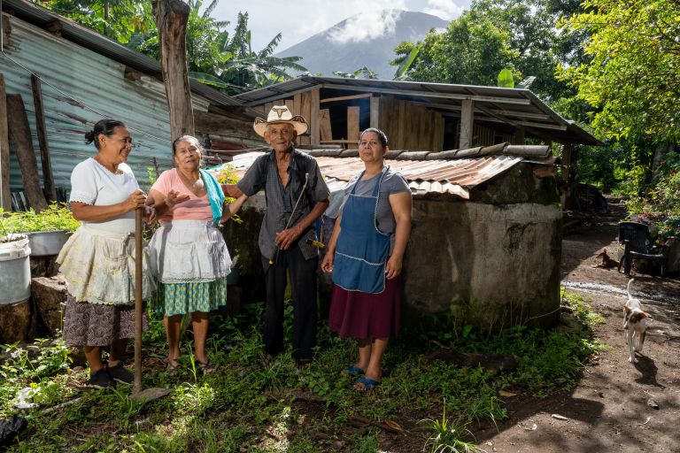 Drei Frauen und ein Mann mit Hut stehen in El Salvador vor einem Haus. Eine Frau hat eine Hacke in der Hand.