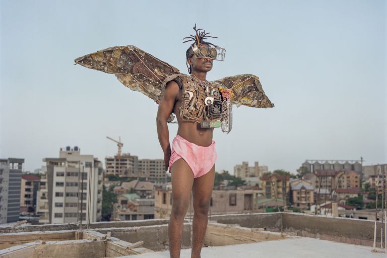 Ein schwarzer Mann steht in rosa Shorts und mit aus Schrott gebauten Flügeln auf einem Dach im Stadtviertel Kitambo